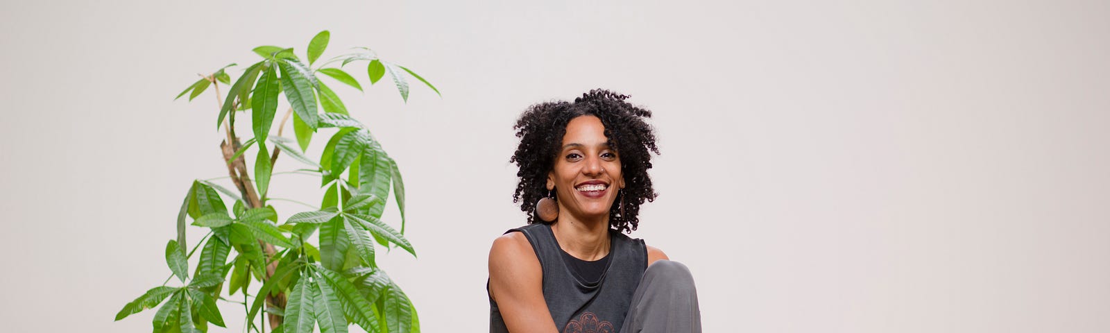 Laia sitting on a wooden stool. She is wearing all gray and is smiling at the camera, sitting next to a luscious Money Tree.