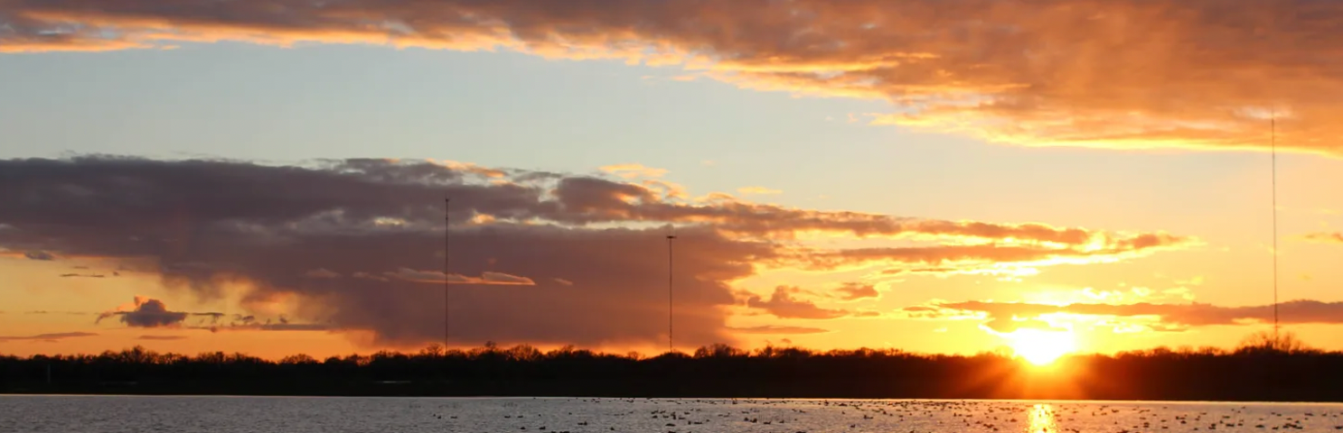 The sun setting in the horizon, with clouds lingering in the air. Sunlight reflects against a body of water in the foreground.