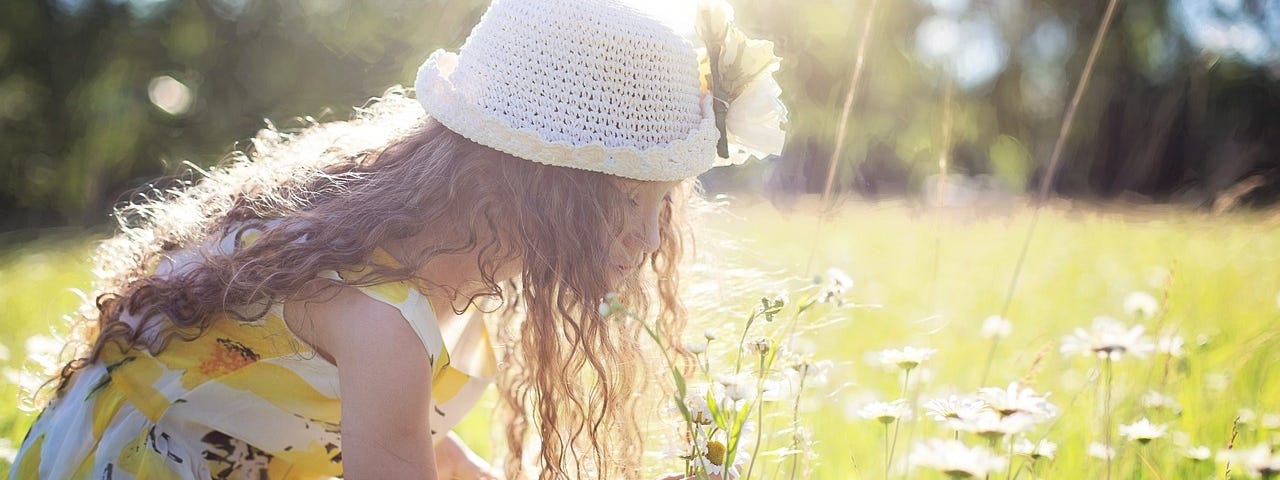 Long haired girl in white crocheted hat and sundress picking daisies as sun begins to set.