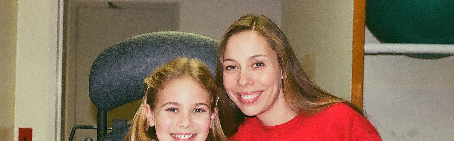 A mom wearing a red sweatshirt with a snowman. The mom has her arm around her ten-year-old daughter who is in a standing frame at the hospital.