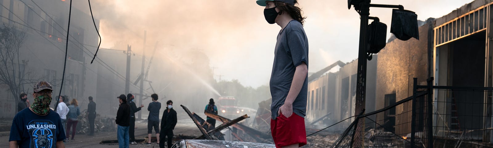 A man stands on a burned out car on Thursday morning as fires burn behind him in the Lake St area of Minneapolis, Minnesota