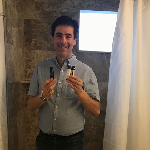 Author holding bottle of shampoo and soap in hotel bathroom in front of shower.