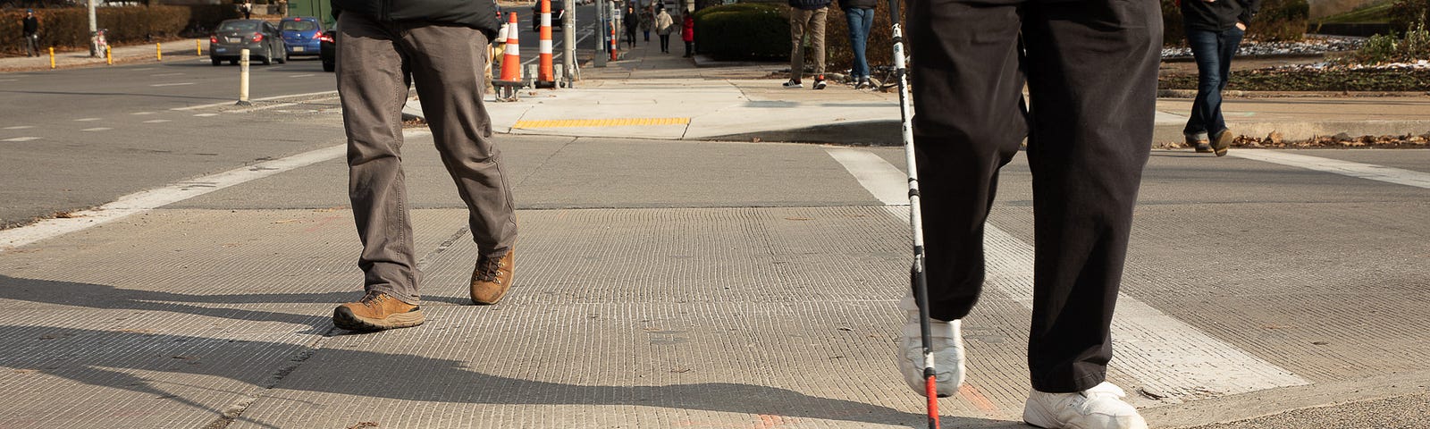 Two people crossing an intersection with the image taken from a low angle showing the curb ramp. One person uses an assistive can typically used by people with visual impairments.