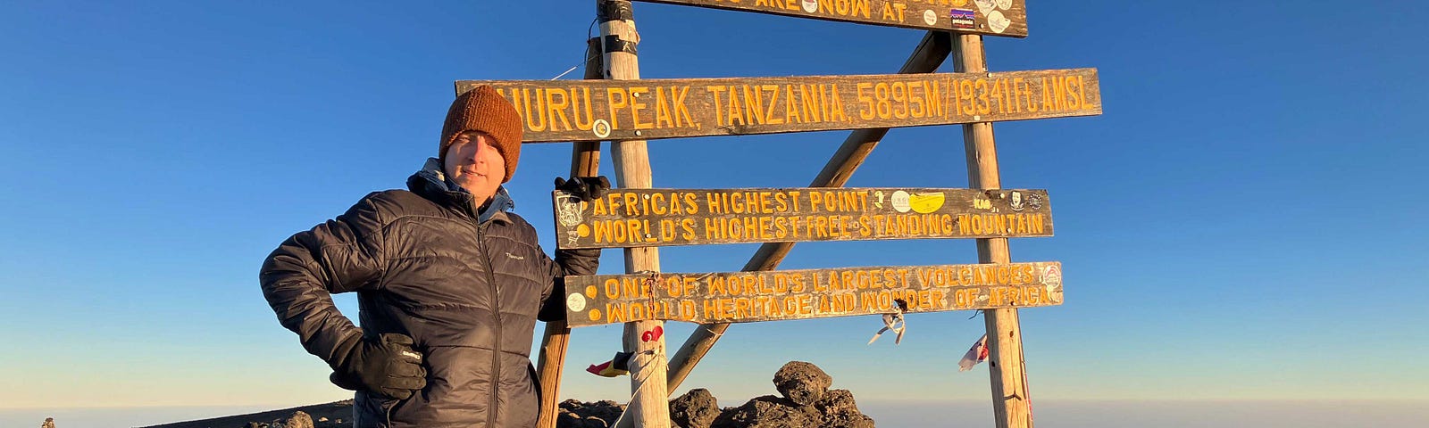 Author standing next to the summit sign at the top of Mount Kilimanjaro at sunrise under deep blue sky.