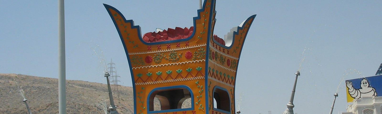 Traffic circle with a tall replica of a traditional Omani incense burner, painted orange with decorations of creepers, surrounded by large silver sprinklers acting as water fountains