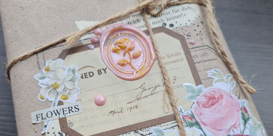 The picture shows a brown package with a collage of paper flowers and print embossed with a pink rose seal tied with brown strings.