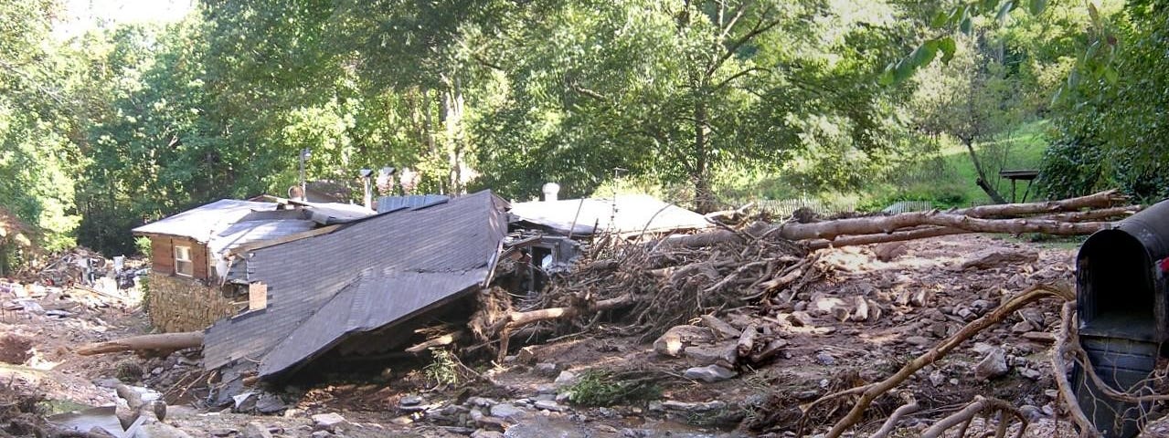 A house with its roof lying detached and boulders, trees, and other debris strewn across the foreground