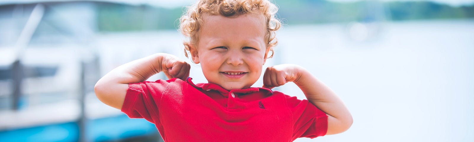 Little boy with blonde curly hair flexing both of his muscles showing his strength in a red shirt.