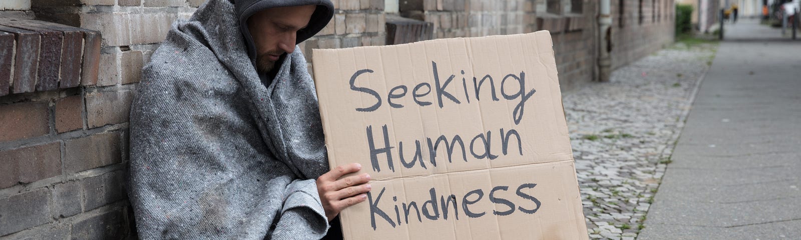 Homeless man sitting on street wrapped in blanket holding a cardboard side that reads Seeking Human Kindness