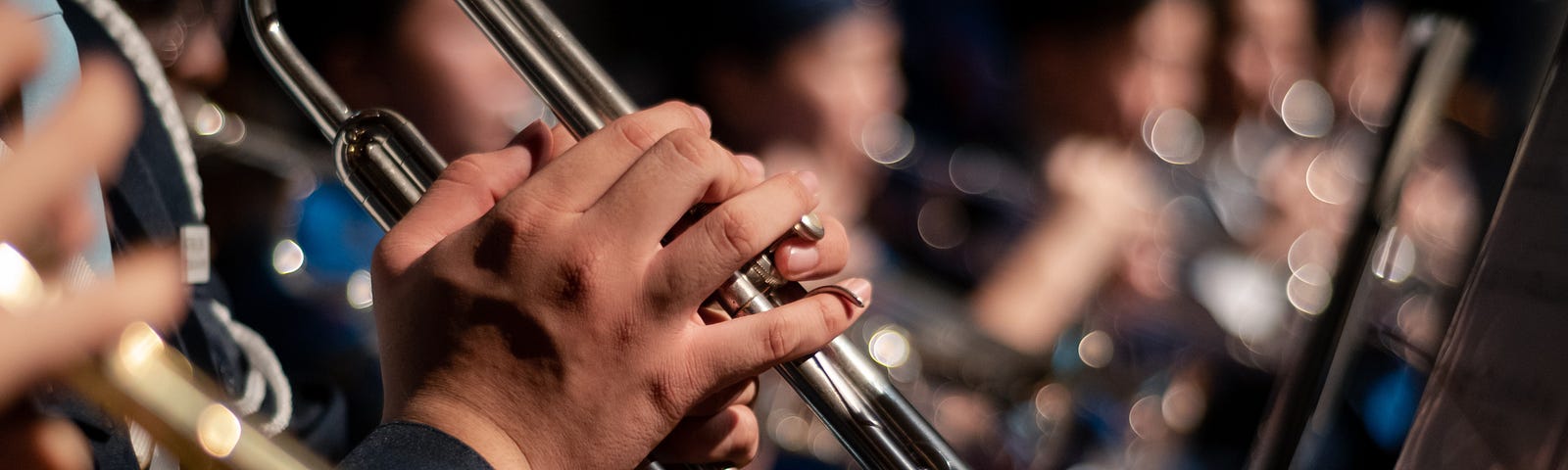 A picture of an orchestral brass section. The photo zooms in on a trumpet player’s hands. In the blurred background, you can see the french horn section.