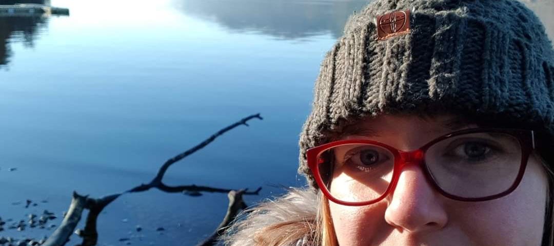 Author of the story smiling with a hat on in the foreground while a still loch is beautiful in sunlight in the background