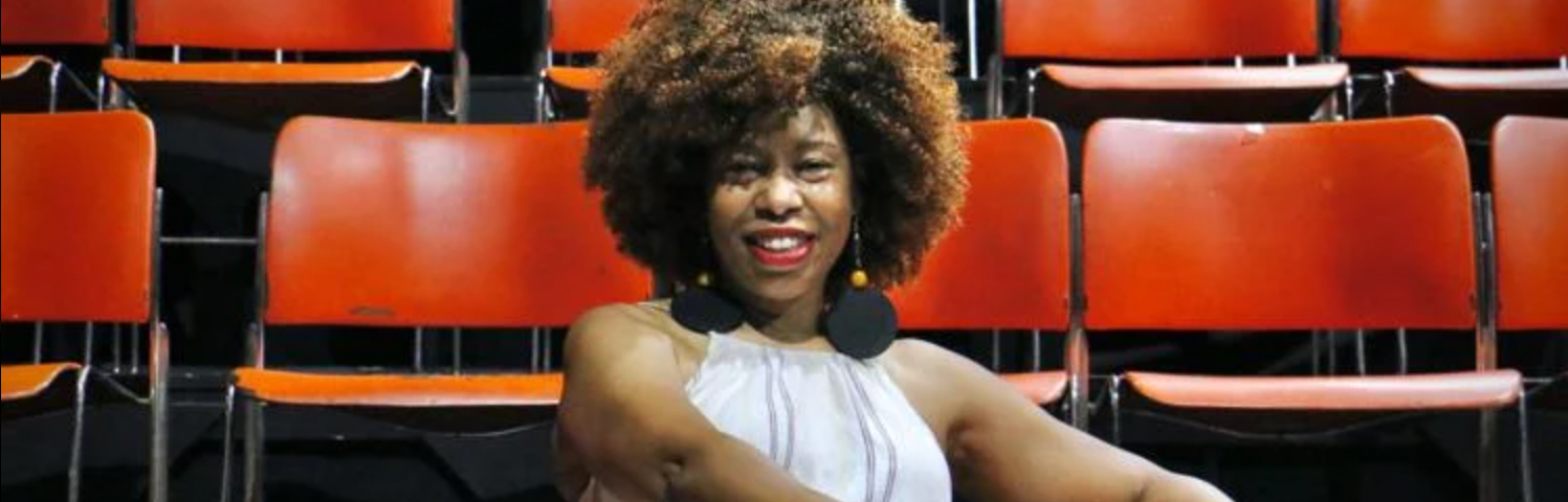 A woman with curly brown hair and brown skin in a white dress sits in a row of orange chairs and smiles at the camera.