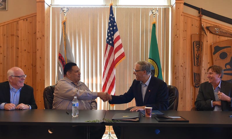 Four men sit at a table with flags behind them representing the United States, Washington state, and Quileute Tribe. Gov. Inslee shakes hands with the man sitting next to him — Doug Woodruff, Chairman of the Quileute Tribal Council — while the other two men look on and smile.