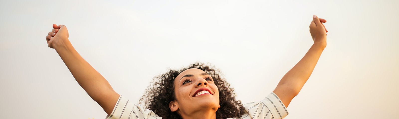 A woman in a sunflower field smiles and holds her arms skyward with positivity.