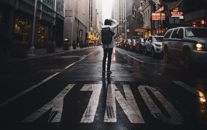 Person standing in middle of Manhattan street, back to camera with right arm raised as if looking for someone.