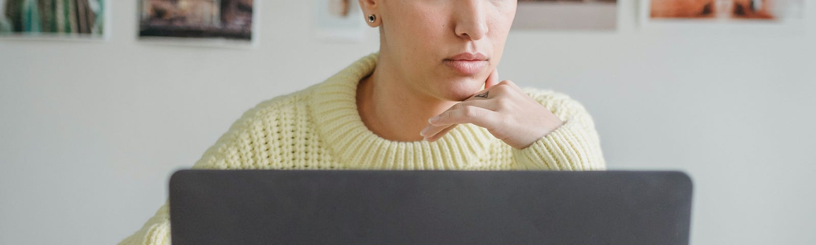 Pensive woman with very short dark hair in yellow sweater using laptop.