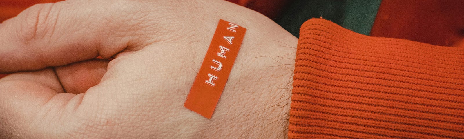 Closeup of a person’s hand with white skin that has an orange sticker on it which reads “human.” The person is wearing an orange coat with a gree shirt underneath.