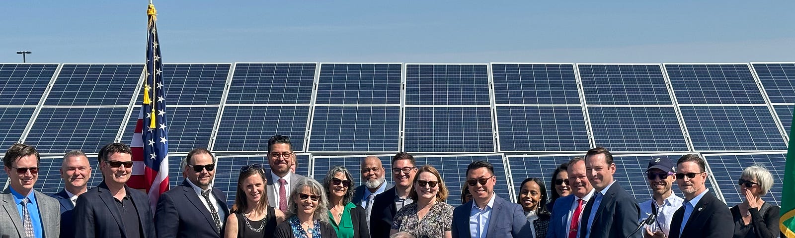 Photo of Gov. Inslee seated at a table signing a bill and a crowd of a couple dozen people standing behind him, smiling at the camera. Behind them are a large array of solar panels and bright blue sky.