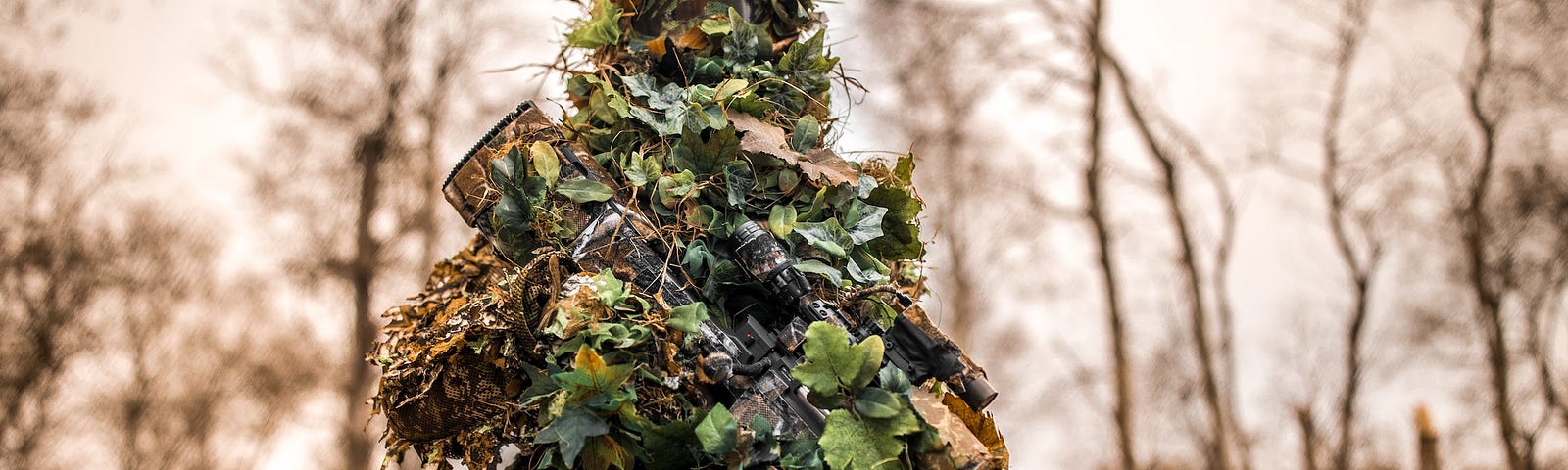 Soldier in ghillie suit holds rifle wrapped in leaves against a forest background, looking ready to shoot UFOs.