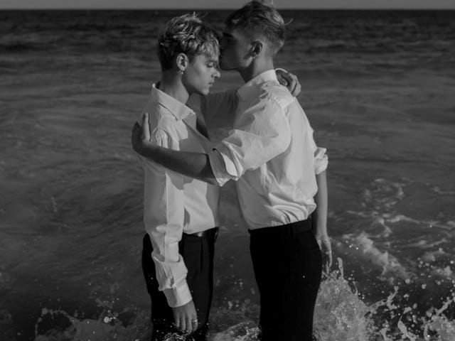 Two men standing in the water with their arms around each other. One man is kissing the other mans head.
