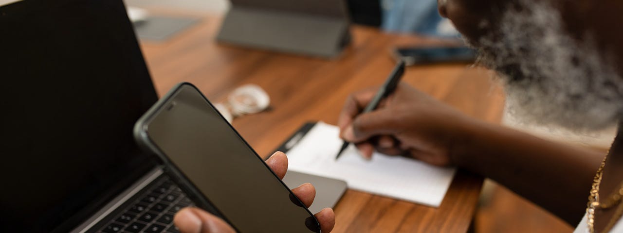 Person of color with a beard, holding a smartphone, writing on paper, at a desk