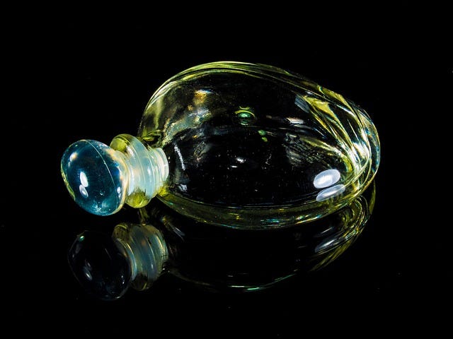A circular corked perfume bottle lying on its side, it’s reflection shown on the table’s surface. Black background