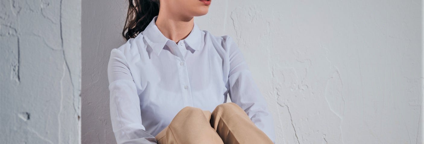 anxious girl with dark hair in ponytail wearing long sleeve shirt and tan pants sitting on floor looking to the side