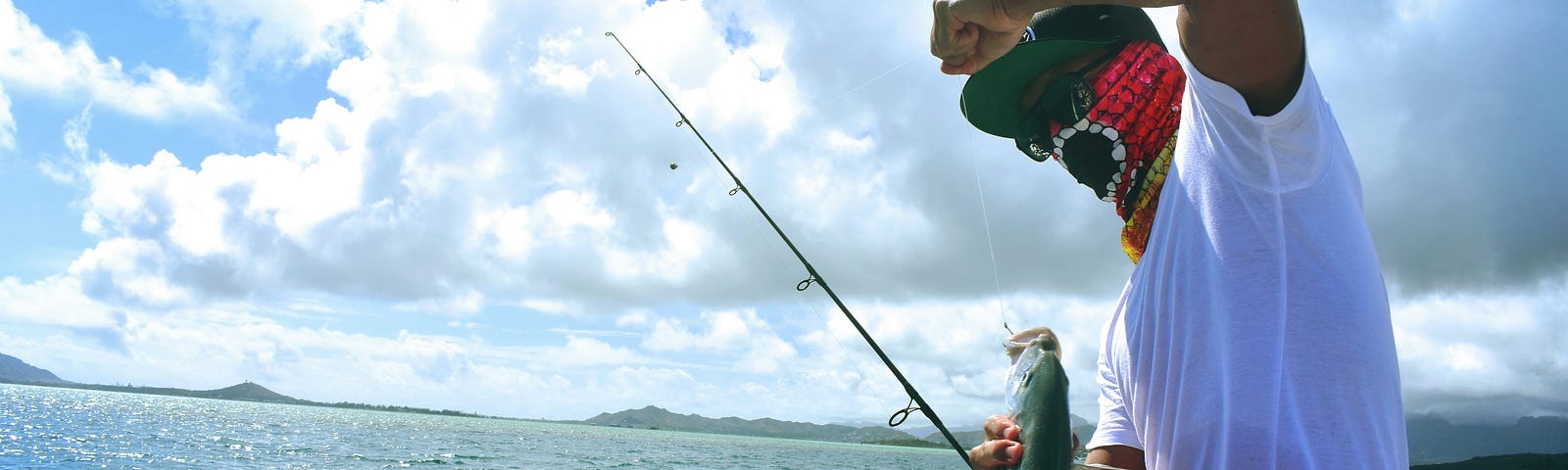 A person in a white shirt and colorful face covering holds a fishing rod over sparkling blue water. They’ve just caught a long, silvery fish that’s dangling in mid-air against a backdrop of distant islands and partly cloudy sky.