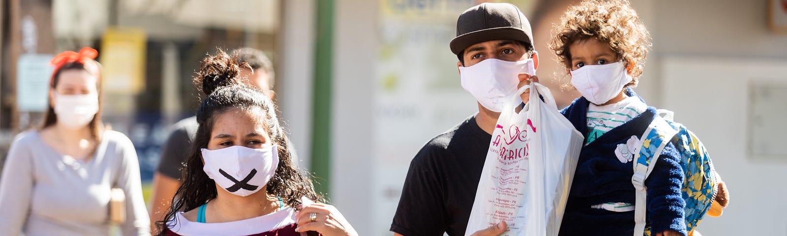 A woman, and a man carrying a young child, walking down the street towards the camera. All three are wearing masks over mouth and nose.