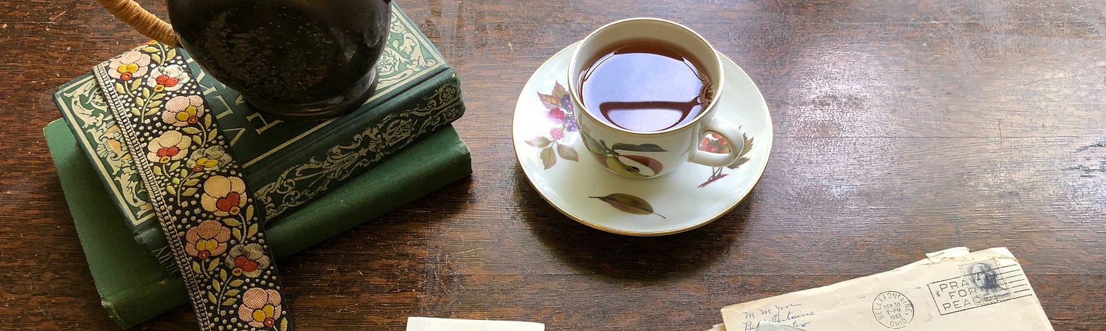 A china teacup and bronze teapot sit on a desk set back from a couple of letters, some books, a piece of paper and a pen.