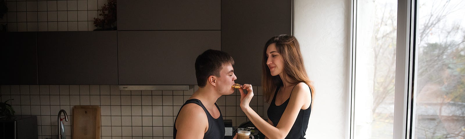 A healthy couple flirtaciously feed one another in a sunny morning kitchen.