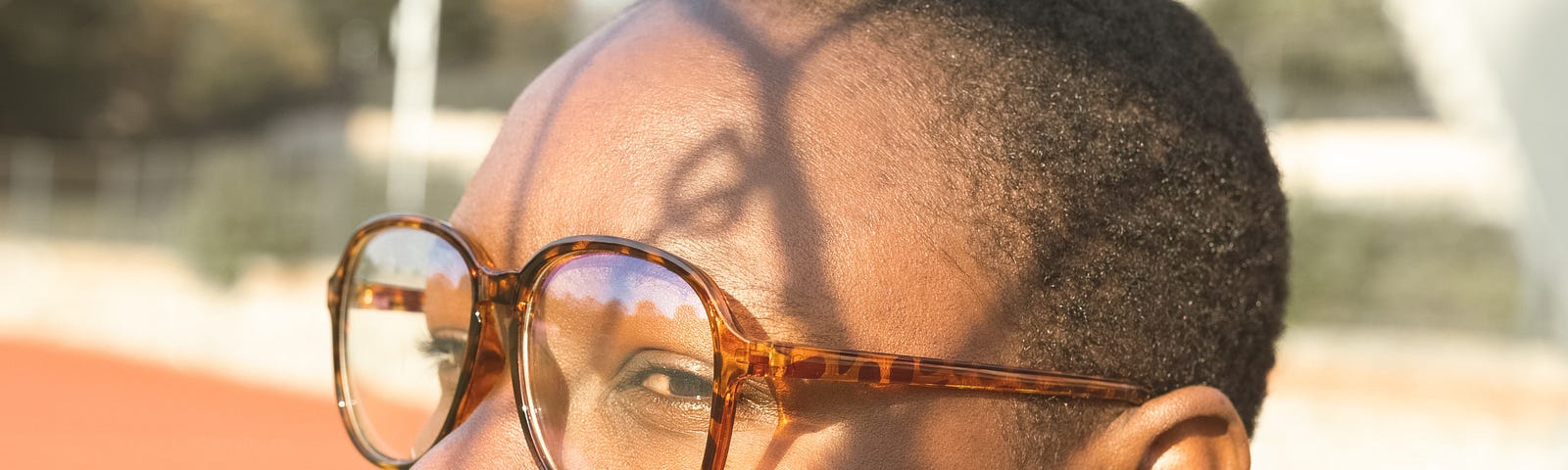 A lady wearing a crew cut and shades. The shadows of some tree branches have lined her face.