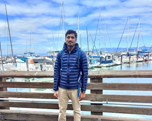 The author stands against a pier with khakis and tennis shoes. There are lots of boats in the background and the sky is beautiful blue.