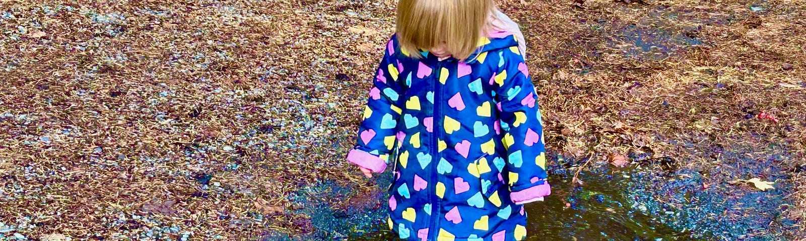 A small child stands in a muddy puddle. She is wearing boots and a rain jacket that is multicoloured.