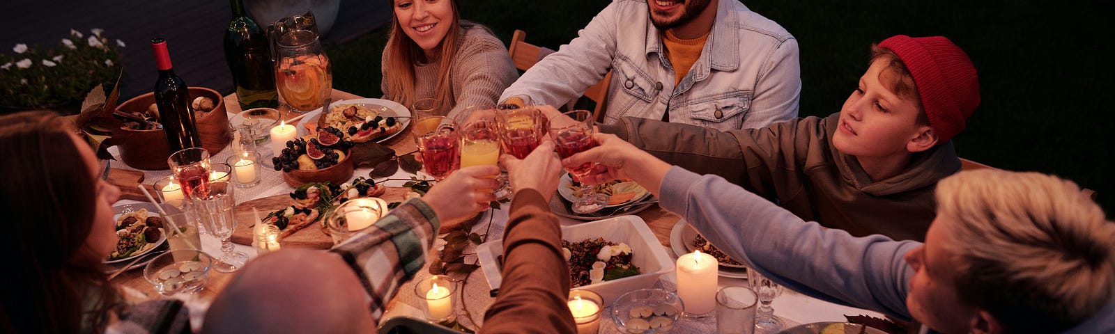 A group of people share a meal and wine around a table in a beautiful atmospheric outdoor setting and enjoy each other’s company as they raise their glasses.