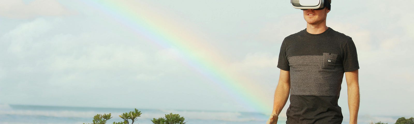 Color photo of a rainbow behind a man walking in a field wearing virtual reality goggles.