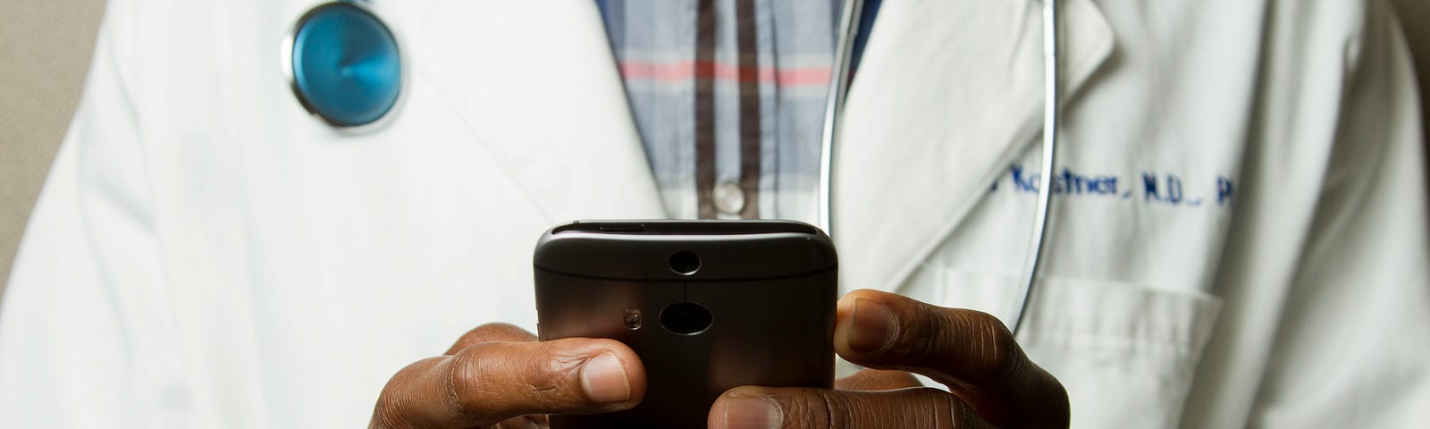 Black Doctor Holding Cell Phone. He has on a plaid shirt under a white coat and has a stethoscope around his neck. His face is out of the frame.