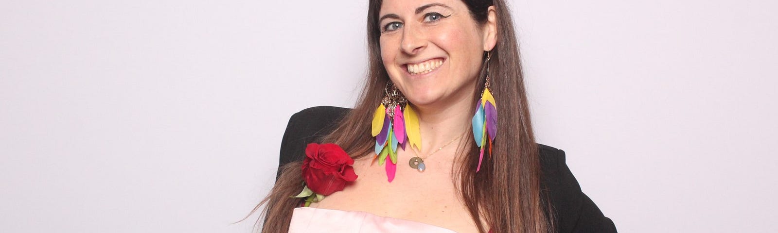 The author, me, dressed in a smart outfit composed of a satin baby pink dress and a black vest complimented with  multicolored feather earrings.
