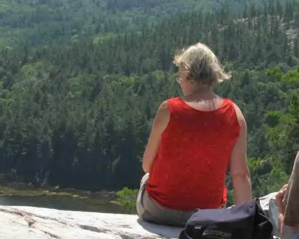 Woman with her back to the camera in a red sleeveless shirt sits on the edge of a cliff looking down at trees.