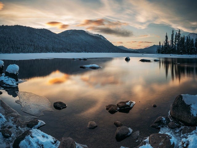 A winter nature scene with mountains and clouds reflected on a lake.