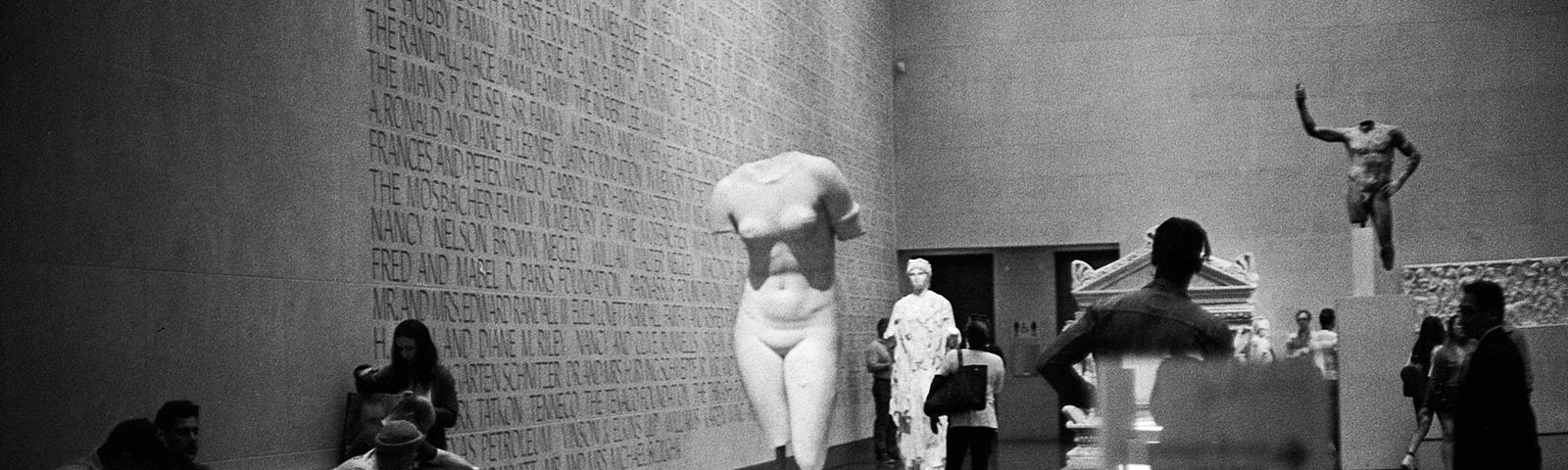 Black and white photo of a museum interior, with a woman’s body statue missing the head, arms, and feet
