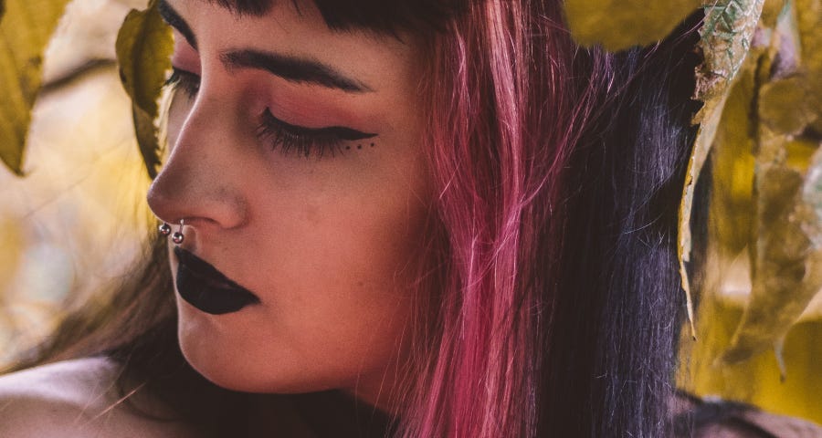 An up-close photograph of a young woman with a nose piercing, black lipstick, and dark hair streaked with red-pink dye.