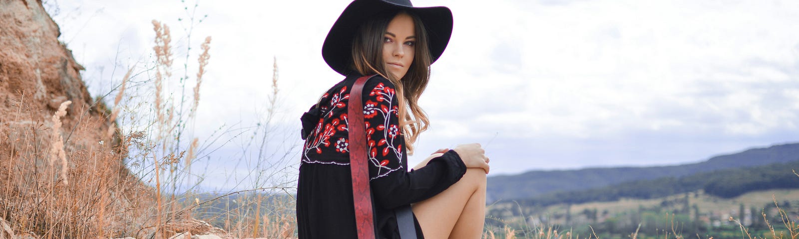 A woman in Western apparel sits in rocky brush amid a mountainous landscape in summertime.