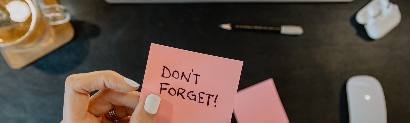 Manicured hand holding a post it reading “Don’t forget” above a computer keyboard and mouse