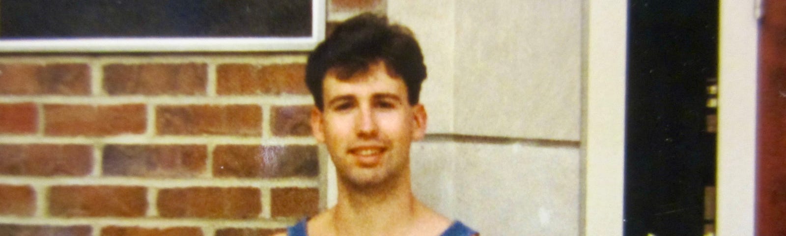 A young man in a blue tank top stands in front of a brick building