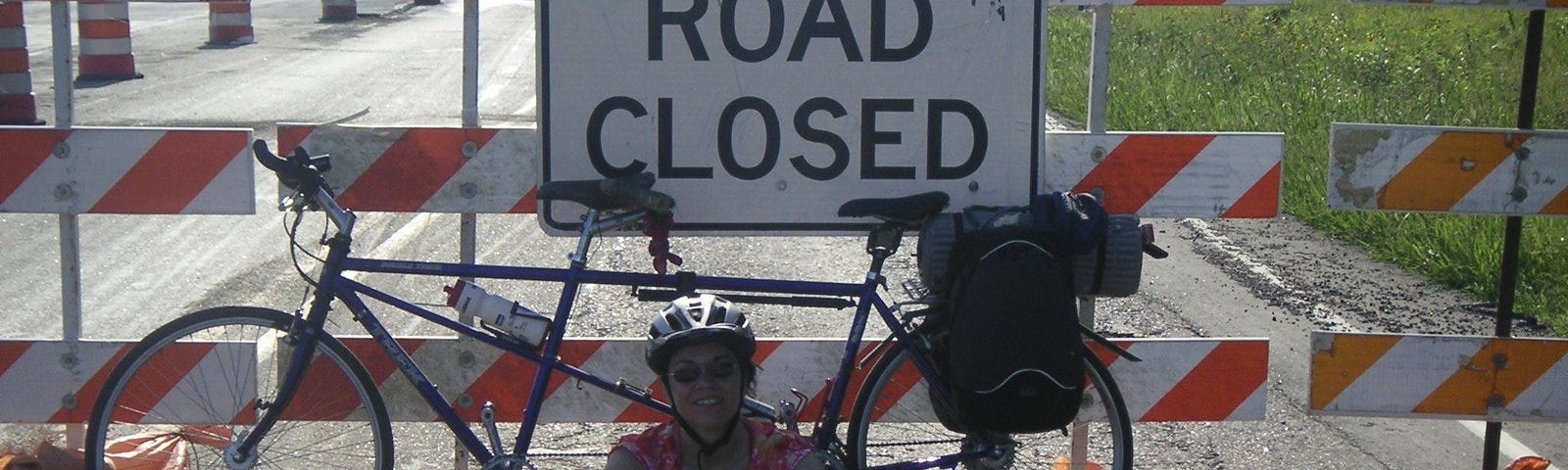 Author, wearing bicycling attire, sits on a paved road in front of her bicycle and a construction barrier which displays a “Road Closed” sign.