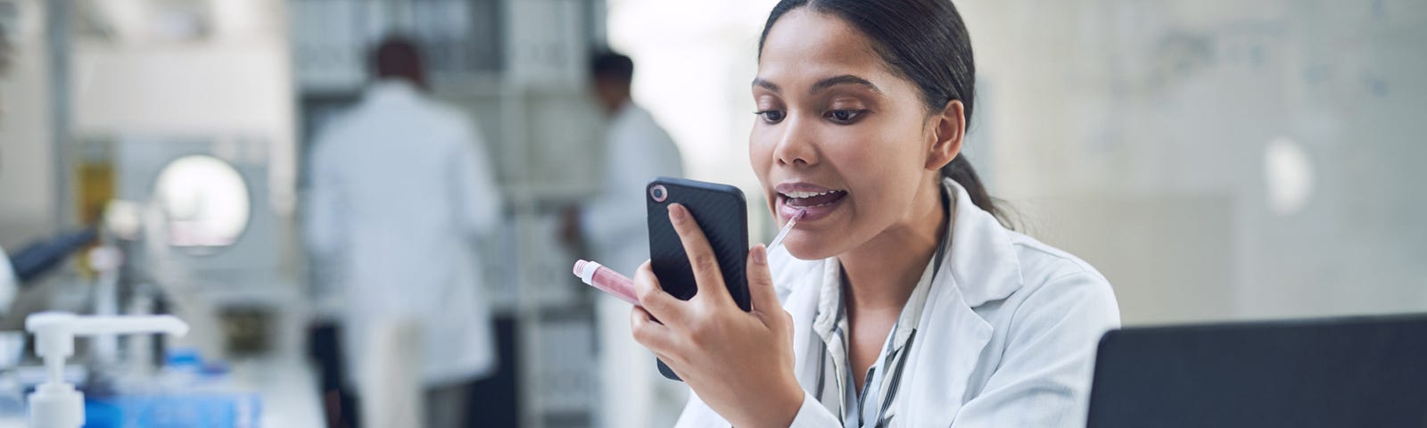 A woman in a lab coat applies lip gloss while staring at her phone. She is at her desk in a laboratory.