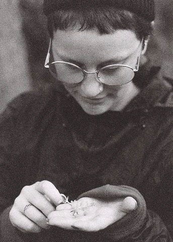 greyscale photo of Charlotte Heather, a white nonbinary person with very short hair and large round glasses. They have a serene facial expression as they look down at a small plant in their hand.
