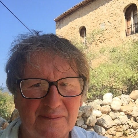 The poet Hugh Macmillan, wearing glasses, in Greece. Behind him — a blue sky, some bushes, a crumbling yellowstone wall and building
