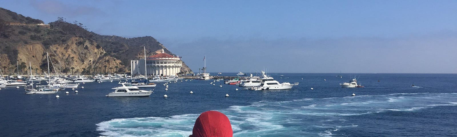 A man in a red baseball cap looks out of the back of a boat to see the beautiful wake as the boat pulls into an island harbor